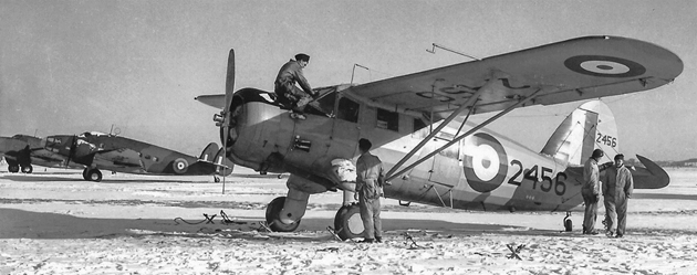 Black-and-white wartime photograph of an RCAF Noorduyn Norseman on a snow-covered airfield, with several airmen standing beside the aircraft and other military aircraft in the background.